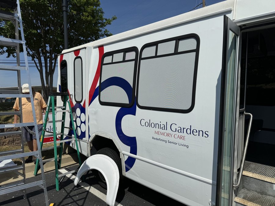 White 'Colonial Gardens Memory Care' shuttle bus with blue-and-red curve motif side wrap