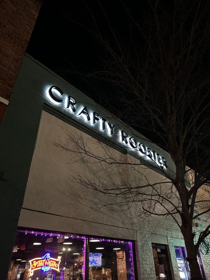 'CRAFTY ROOSTER' dimensional letters illuminated on a dark storefront facade at night