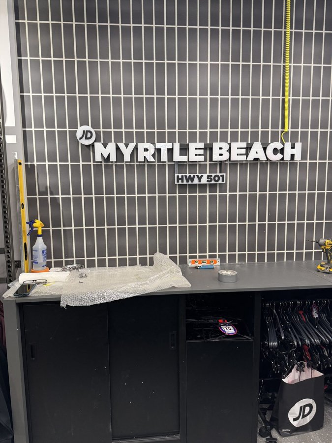 Freshly installed 'JD MYRTLE BEACH HWY 501' dimensional letters on a retail slatwall, install tools on the counter